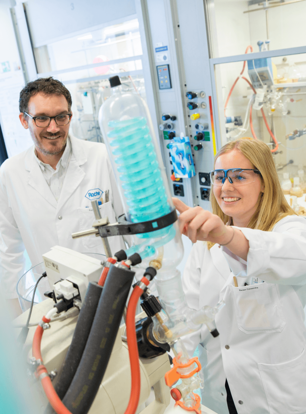 Two smiling researchers in lab coats, a man and a woman, observe a large glass apparatus with a blue spiral structure, the woman pointing at it.