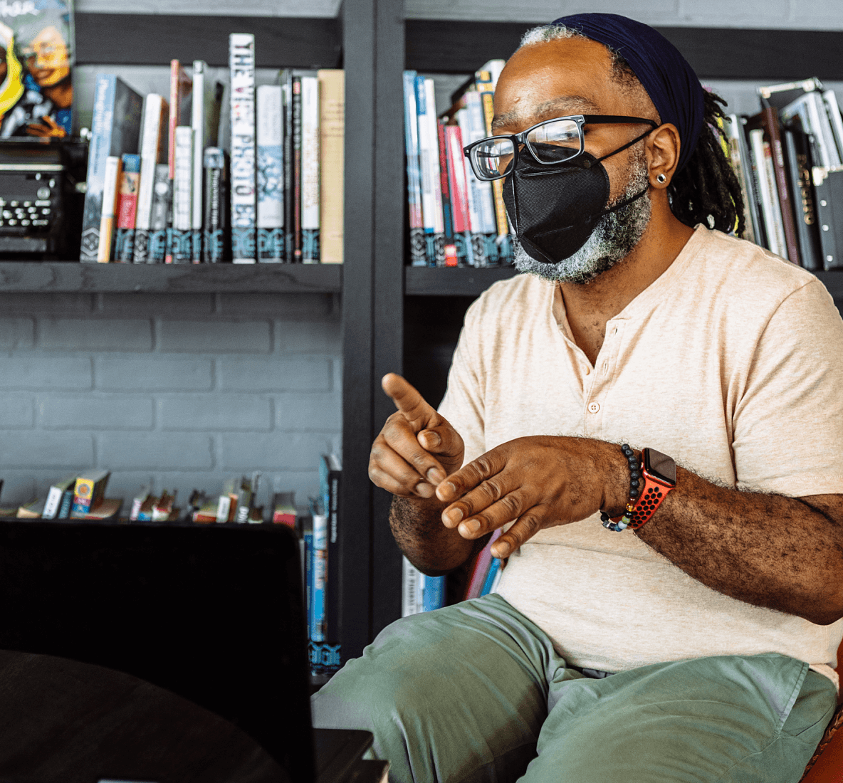 Portrait d'un homme noir sourd portant des lunettes et un masque KN-95, signant lors d'une conversation vidéo via un ordinateur portable. L’homme est assis à une table basse dans une bibliothèque municipale remplie de livres et d’affiches.