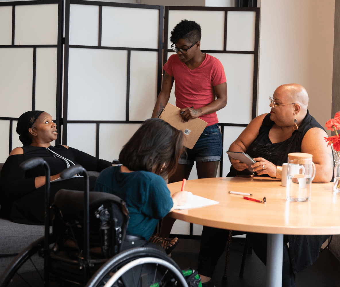 Quatre personnes handicapées racisées sont réunies autour d’une table lors d’une réunion. Une femme noire assise sur un canapé parle avec une expression neutre tandis que les trois autres (une personne sud-asiatique assise dans un fauteuil roulant et prenant des notes, une personne noire non binaire assise sur une chaise avec une tablette et une canne, et une personne noire non binaire debout avec un bloc-notes) écoutent.