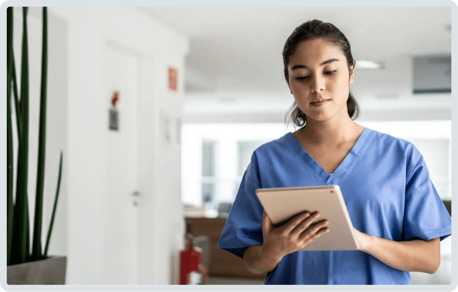 Woman in blue scrubs looking at a tablet in a hallway.