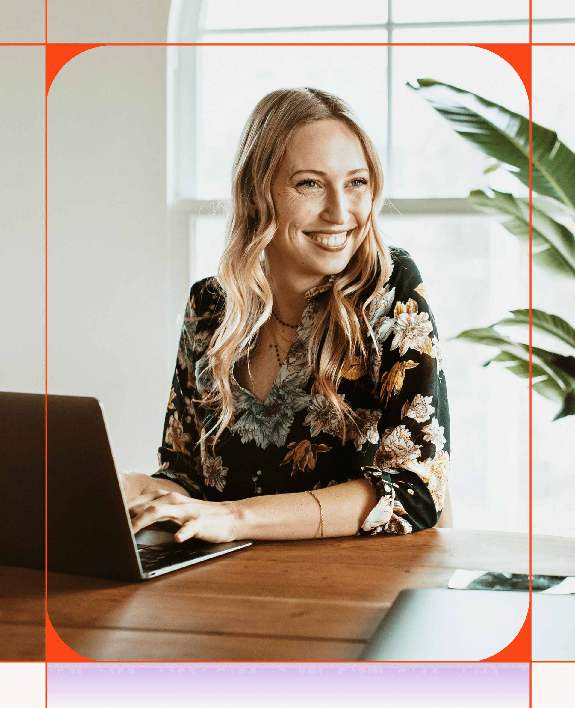 A smiling blonde woman in a floral blouse types on a laptop at a wooden table.