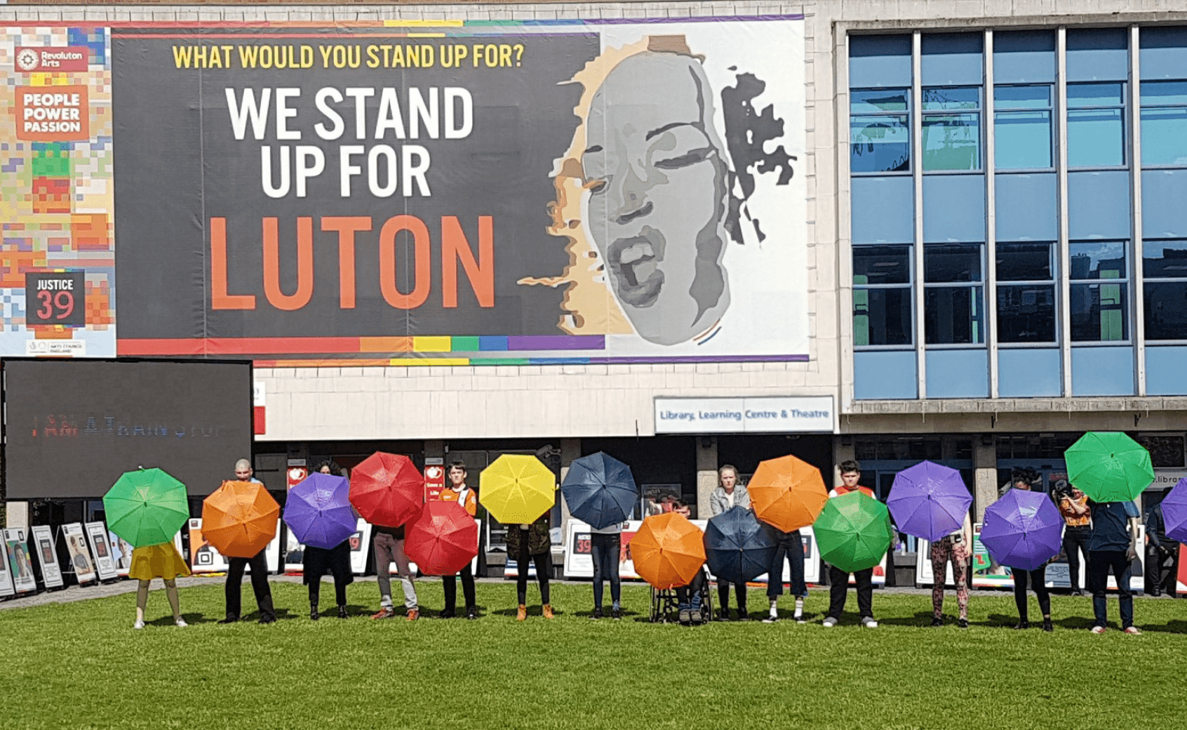 A line of people stands on grass holding brightly colored umbrellas forming a rainbow, in front of a building with a large banner reading "WE STAND UP FOR LUTON" and a grayscale face.