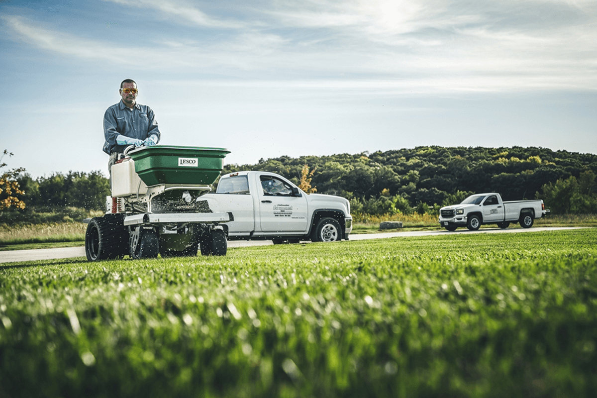 A man operates a walk-behind fertilizer spreader on a large green lawn, with two white pickup trucks and trees in the background.