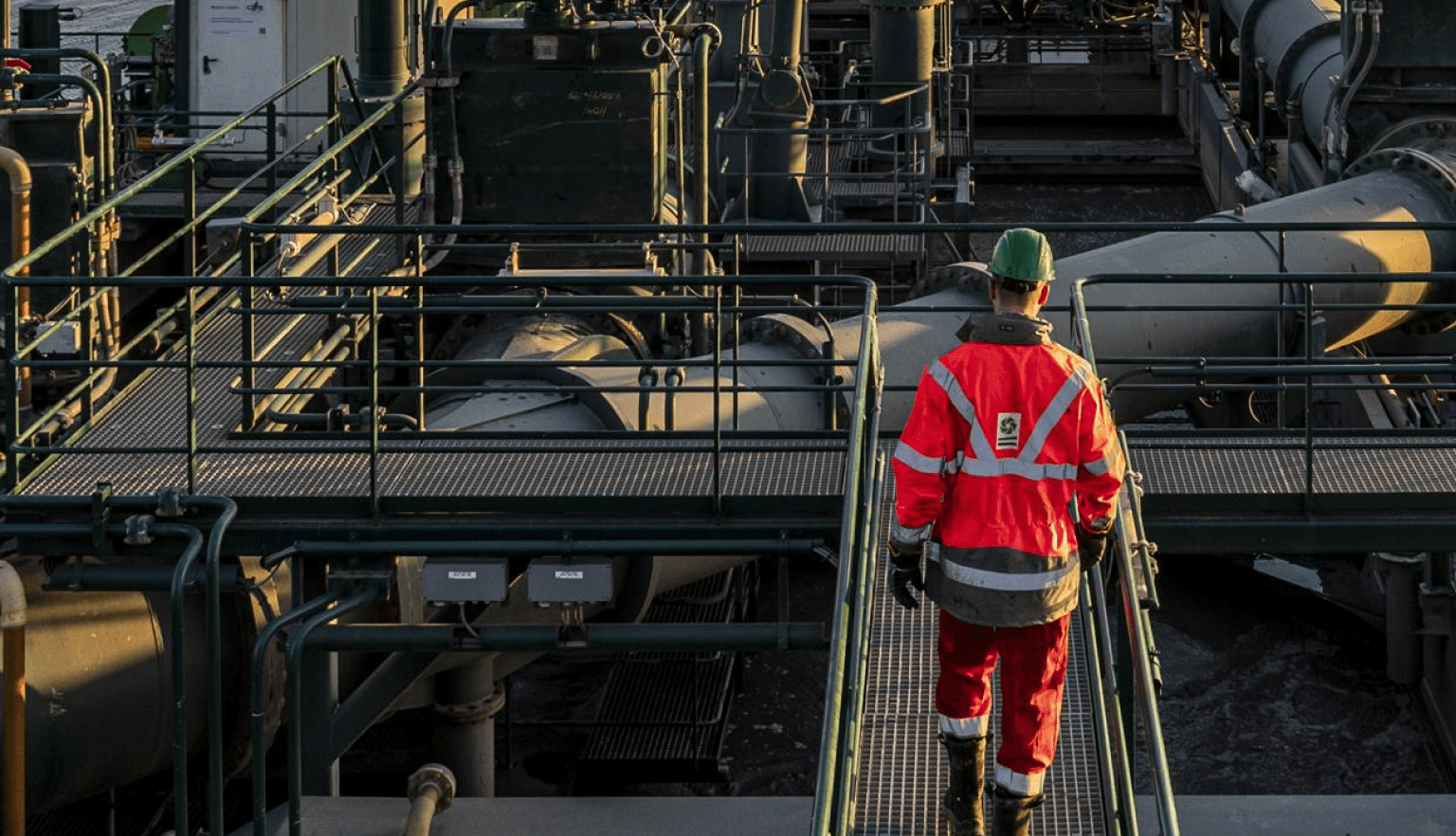 Worker in safety gear walks on a metal platform through an industrial facility.