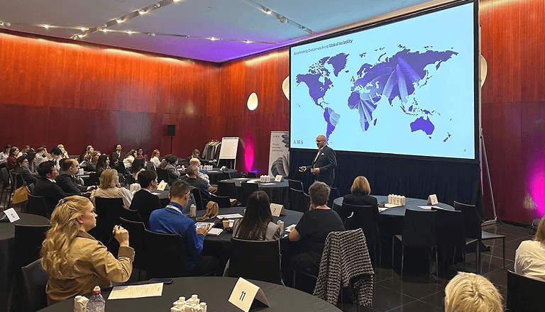 A speaker presents a world map with data on a large screen to an audience seated at tables in a conference room.
