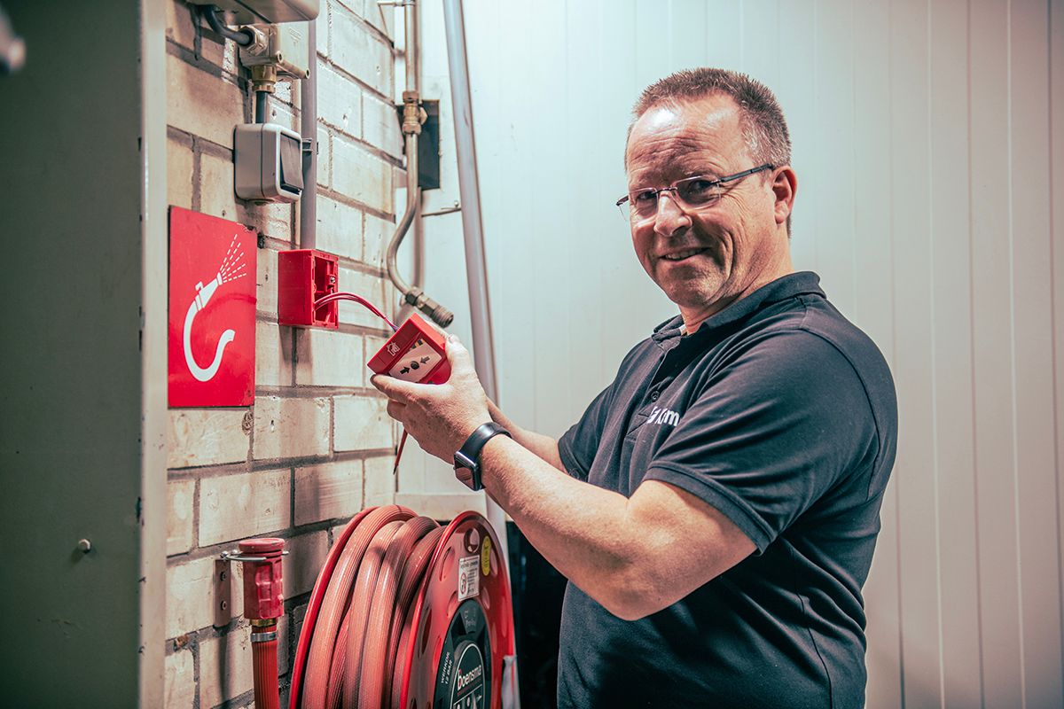 A smiling man in glasses works on a red fire alarm next to a fire hose reel.