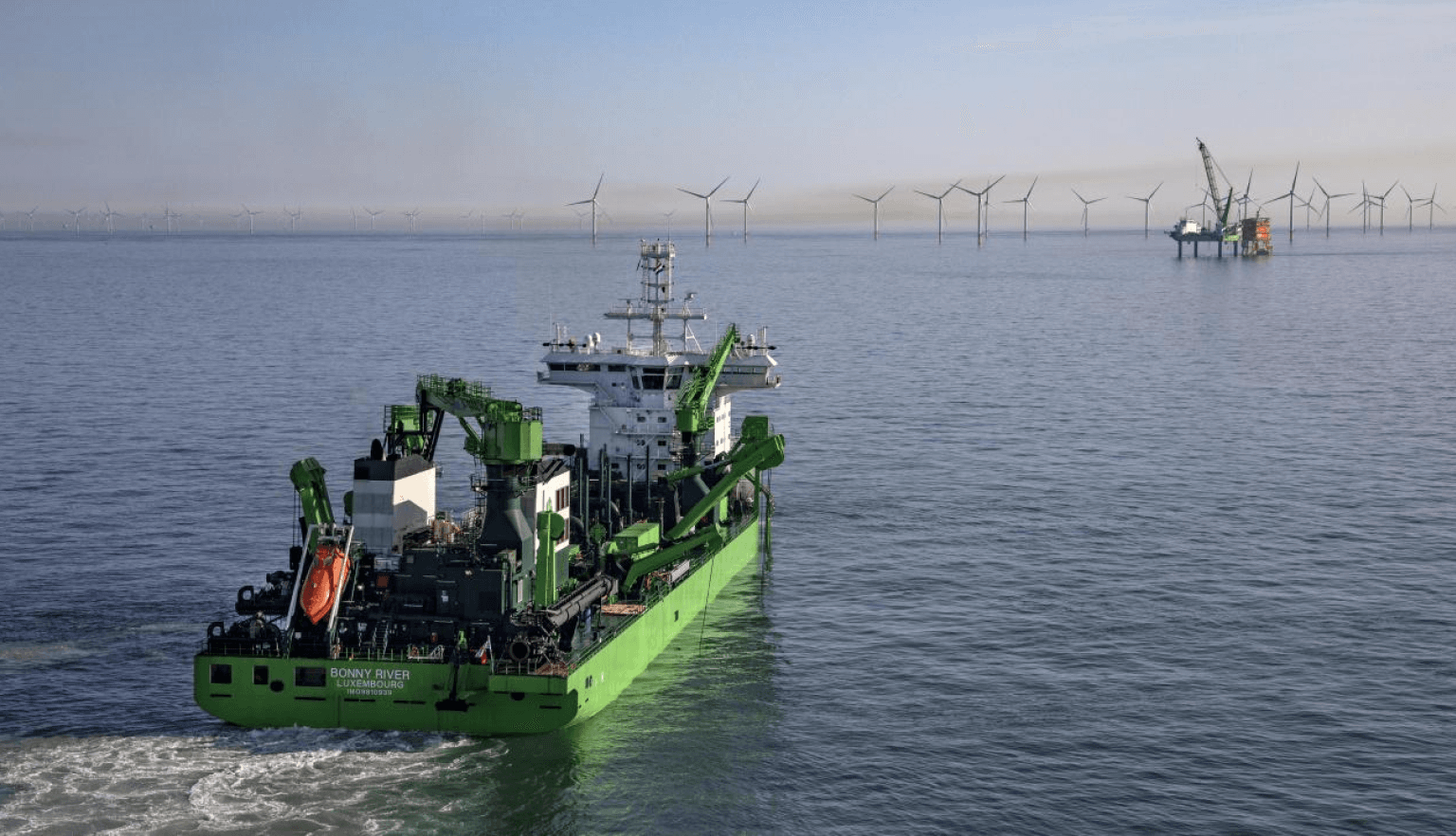 A large green offshore vessel sails in the ocean, with an offshore wind farm and a crane platform visible in the distance.
