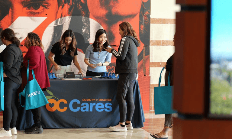 Donne interagiscono presso uno stand all'evento "County of Orange OC Cares", alcune con borse di tela con la scritta "HIRE".