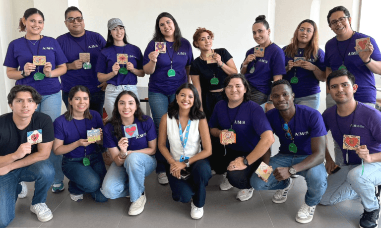 Un gruppo eterogeneo di persone sorridenti, molte delle quali con indosso magliette viola A.M.S., che mostrano piccole carte decorate.