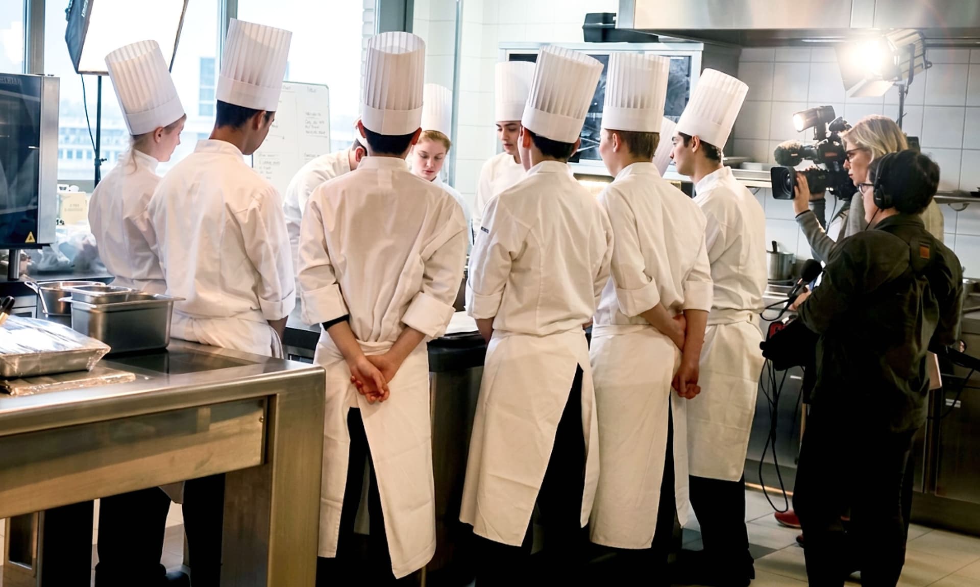 Chefs in a professional kitchen being filmed by a camera crew.