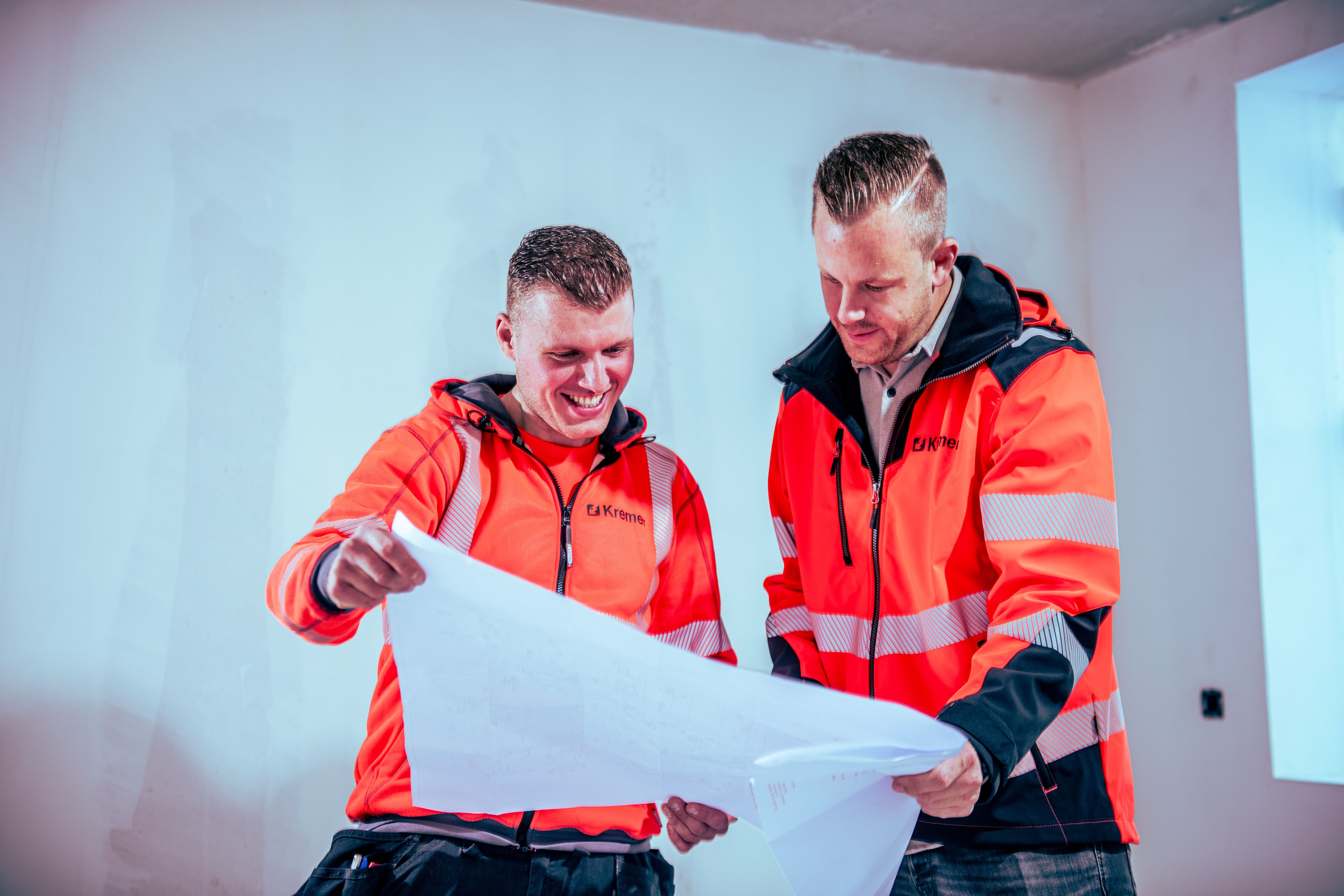 Two construction workers in orange high-visibility jackets smile while reviewing blueprints in an unfinished room.