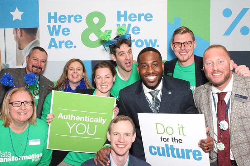 A group of nine smiling people, some in green attire, hold signs that read "Authentically YOU" and "Do it for the culture" in front of a "Here We Are & Here We Grow" backdrop.