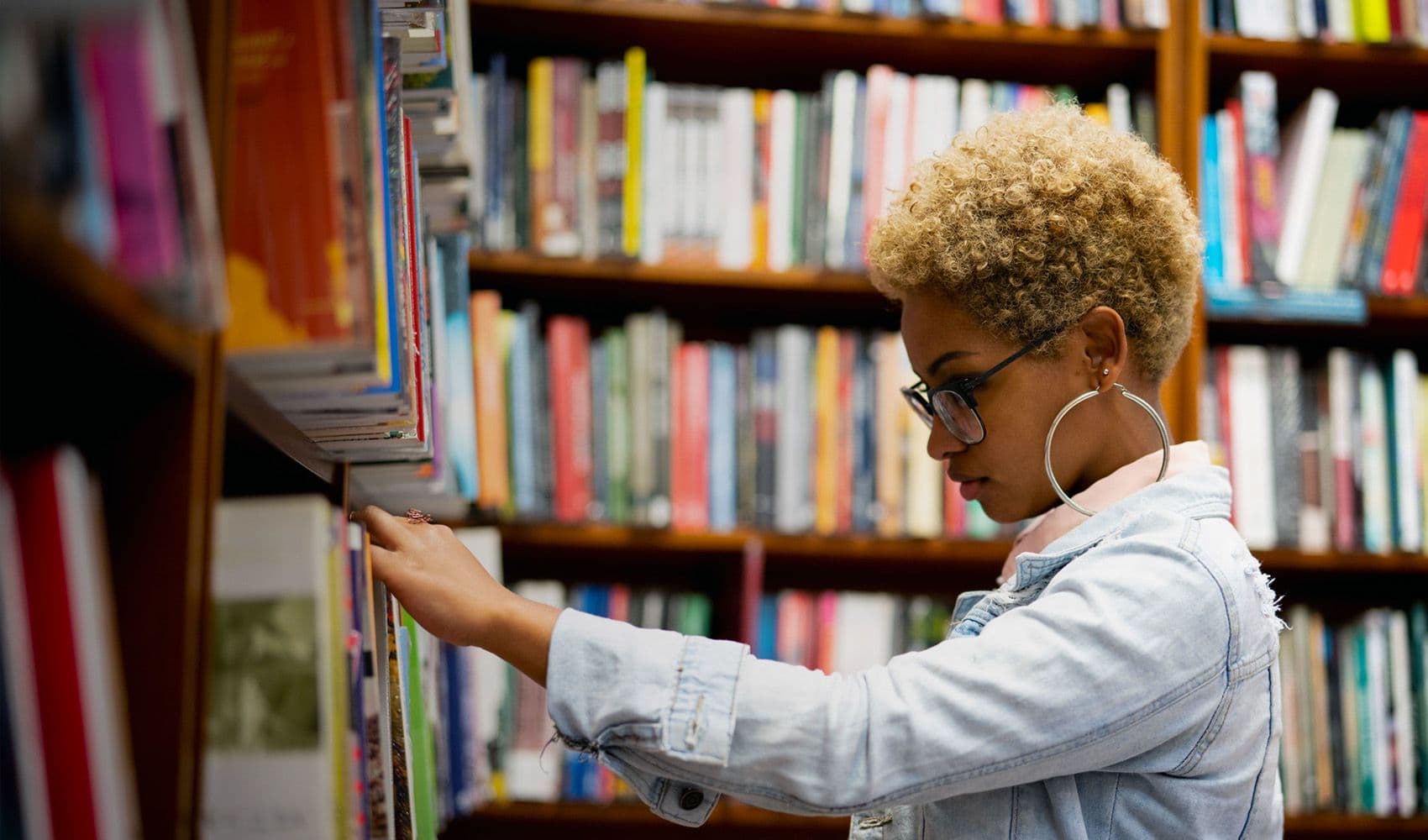 Young Black woman with blonde afro, glasses, and hoop earrings browsing books on a shelf.