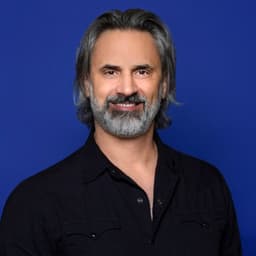 Smiling man with gray hair and beard in a black shirt against a blue background.