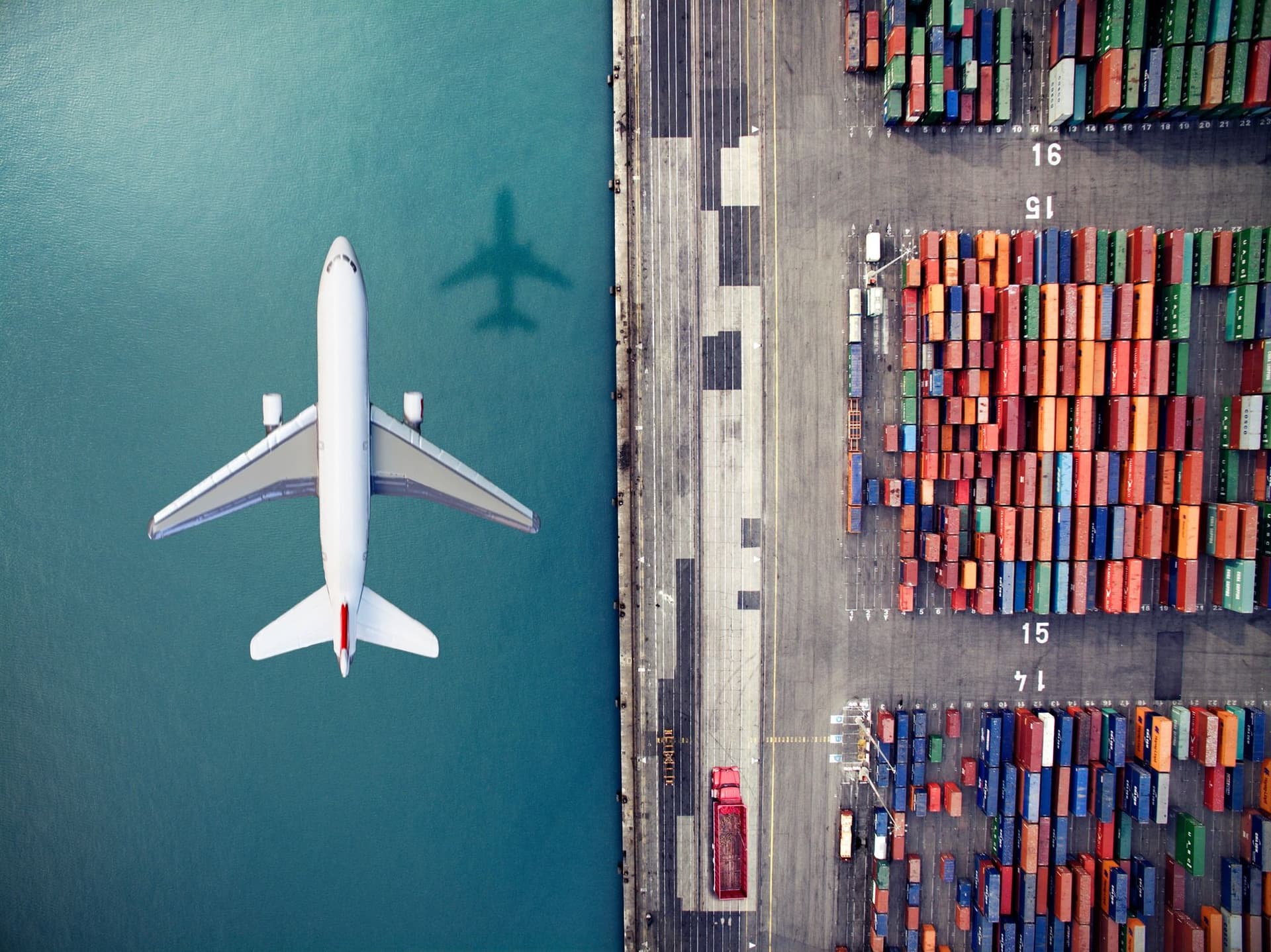 Aerial view of a white airplane flying over water next to a port filled with colorful shipping containers.