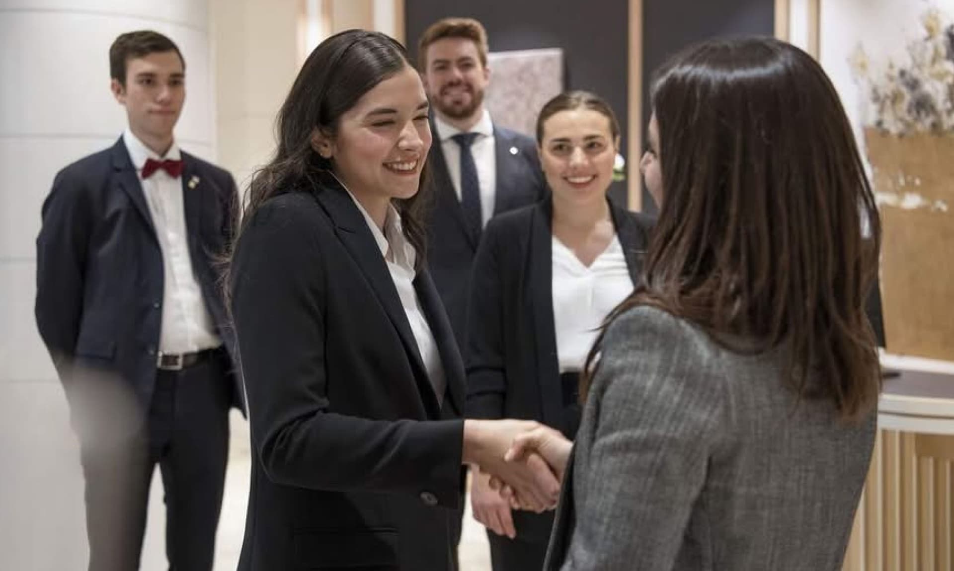 Two women in business attire shake hands, with three colleagues standing in the background.