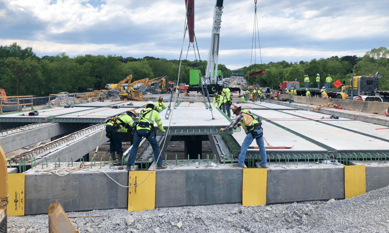 Construction workers use a crane to install concrete slabs for a new bridge or overpass, with other heavy machinery and traffic visible in the distance.