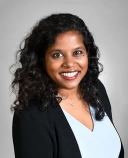 Smiling woman with dark, curly hair wearing a light blue top and black cardigan.