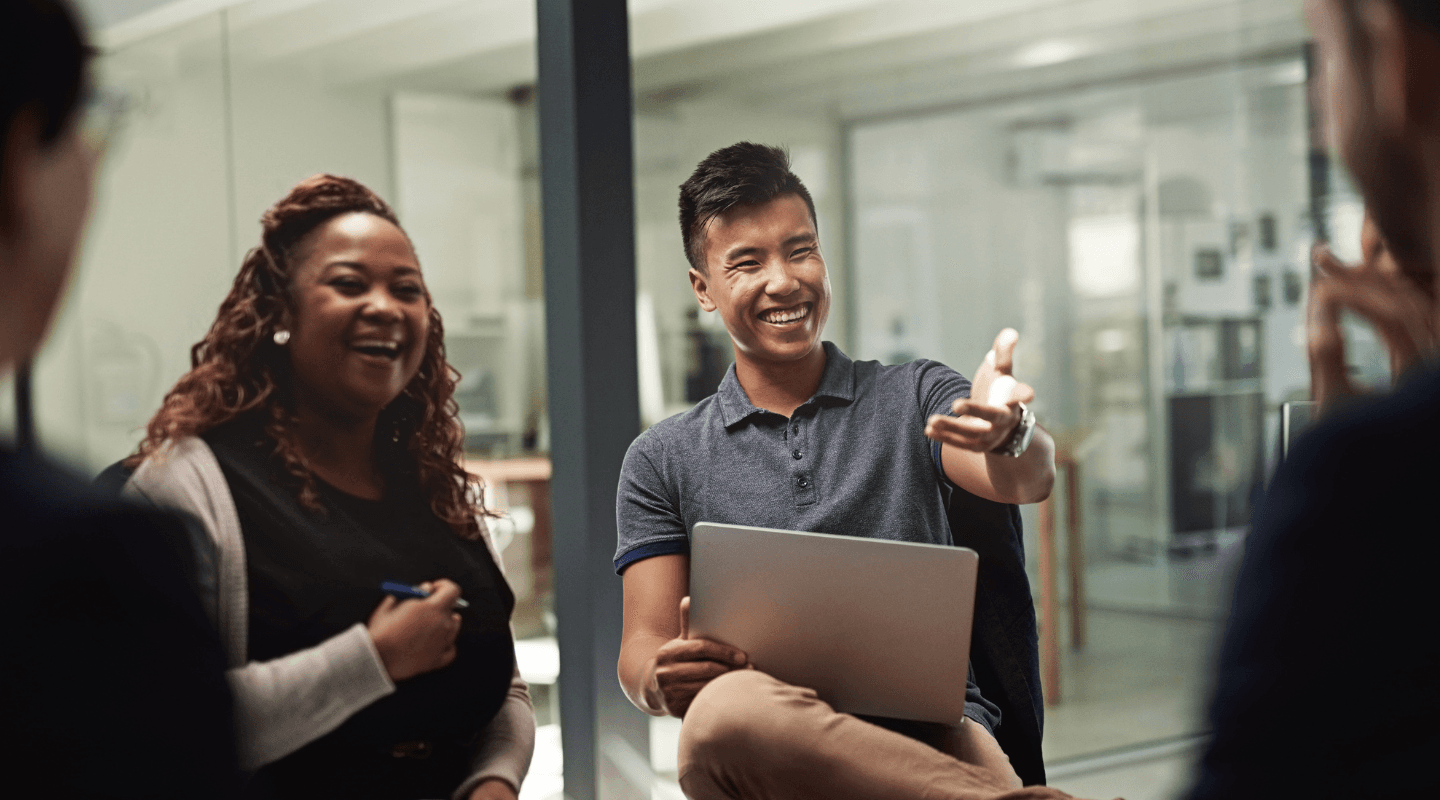 A smiling man holds a laptop and gestures during a diverse team meeting, with a laughing woman beside him.