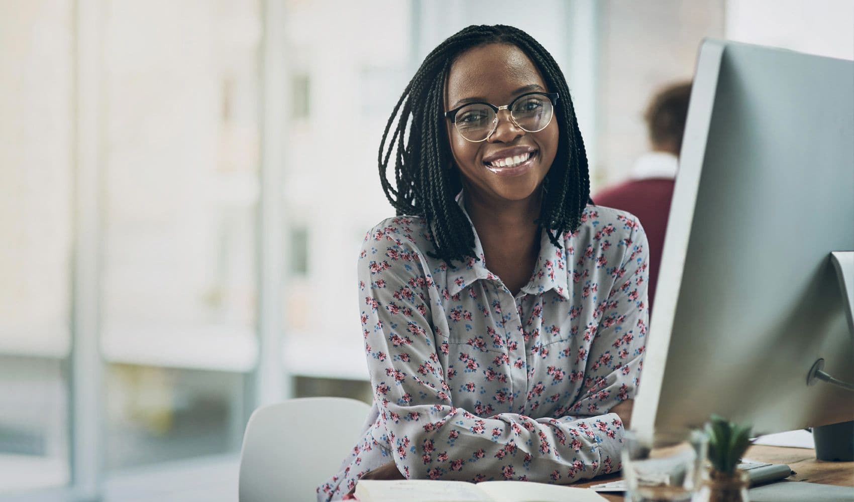 A smiling Black woman with braided hair and glasses sits at a desk in an office, facing forward with a computer monitor to her right.