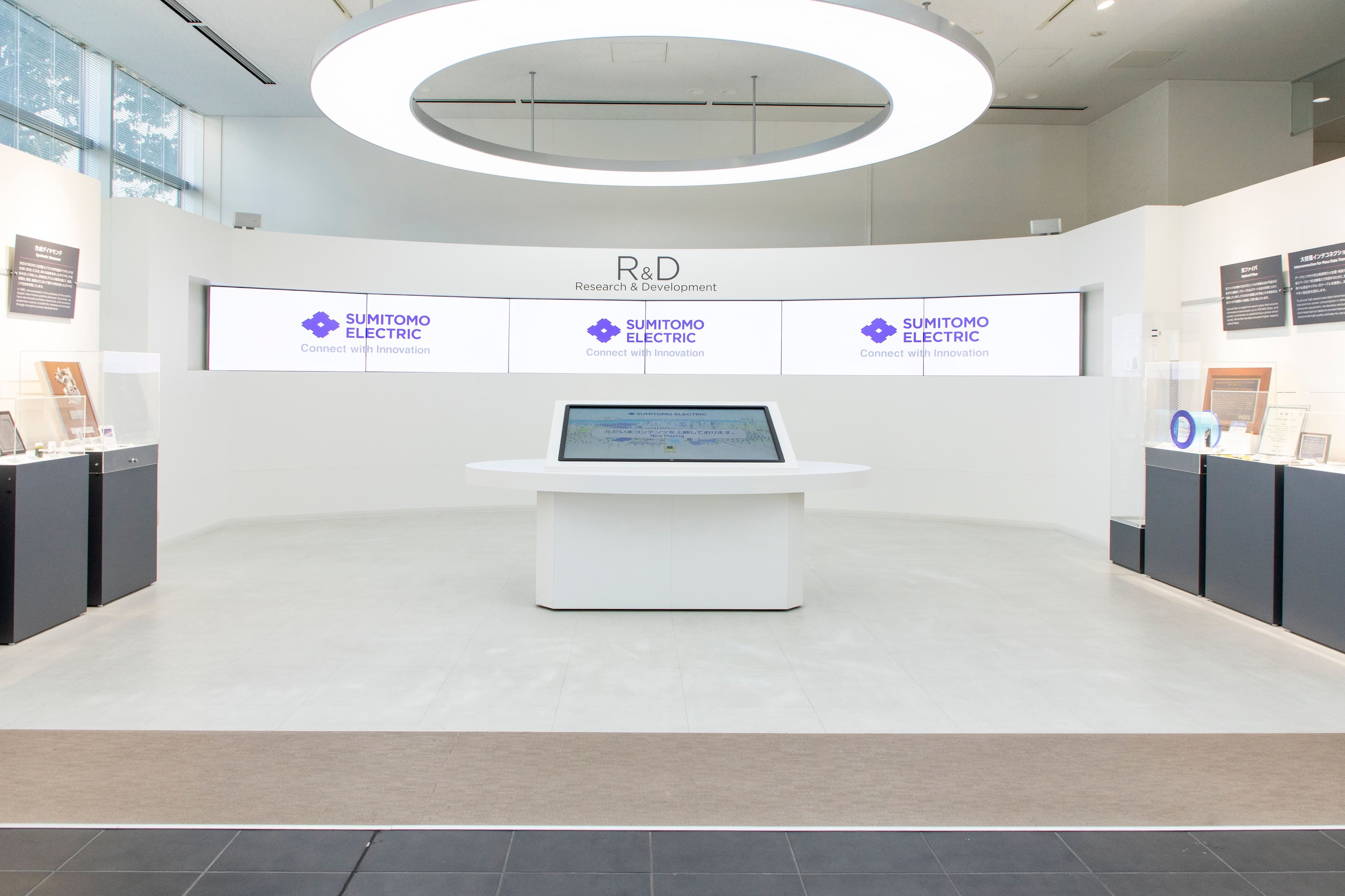 Modern Sumitomo Electric R&D exhibit featuring branded screens, an interactive table, and product displays.