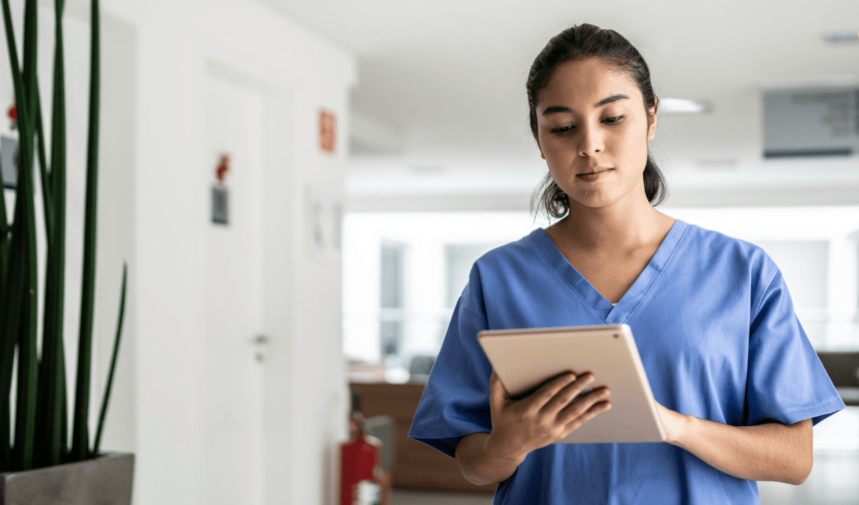 A woman in blue scrubs looks at a tablet in a medical hallway.