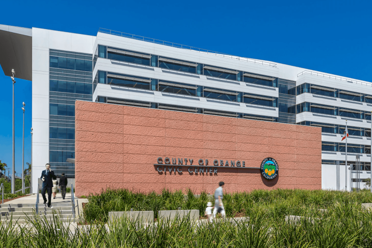 A modern white building, the County of Orange Civic Center, with a large reddish-brown sign wall in front, flanked by green landscaping and blurred people walking.