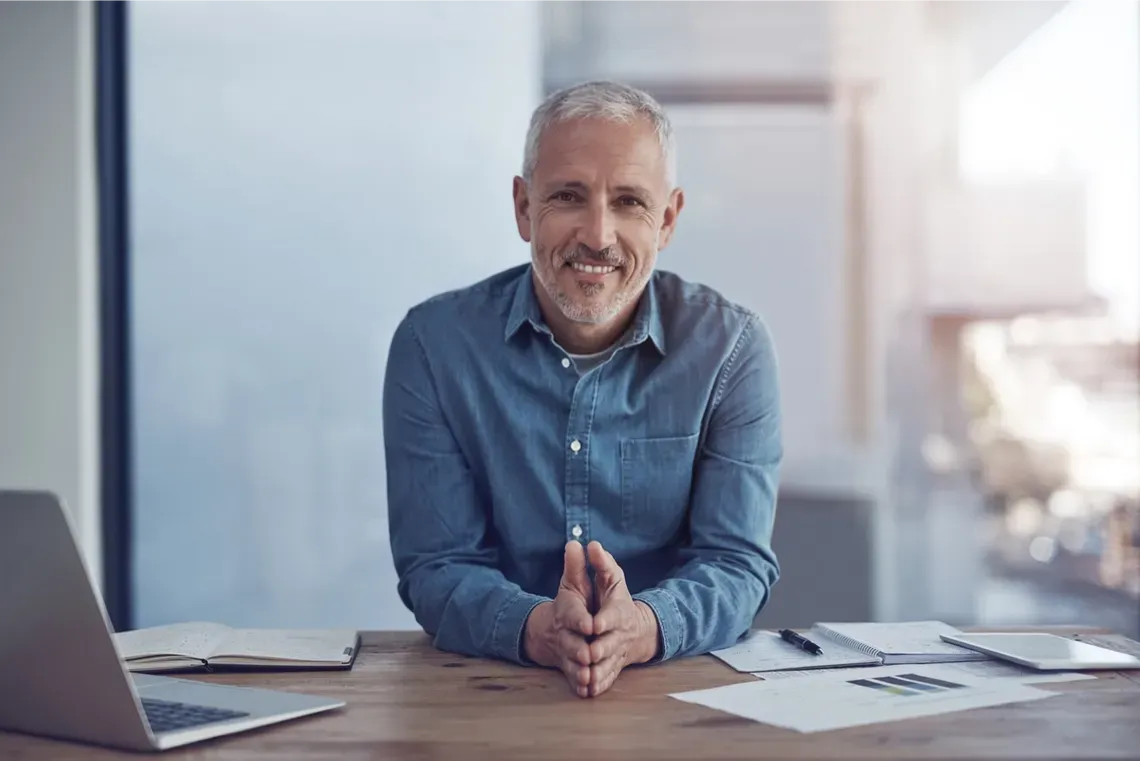 A man is sitting at a desk with his hands folded and smiling.